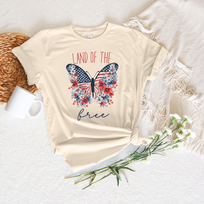 May include: A beige t-shirt featuring a butterfly design. The butterfly has the American flag on its wings and is surrounded by red, white and blue flowers. The text "Land of the free" is printed below the butterfly.