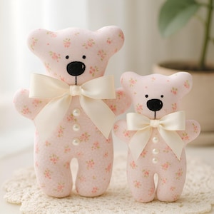 Puede incluir: Dos osos de peluche de tela rosa con estampado floral. Los osos tienen ojos de botón negros, narices negras y lazos color crema. Están adornados con botones de perlas y son de diferentes tamaños, perfectos para una guardería o como regalo.