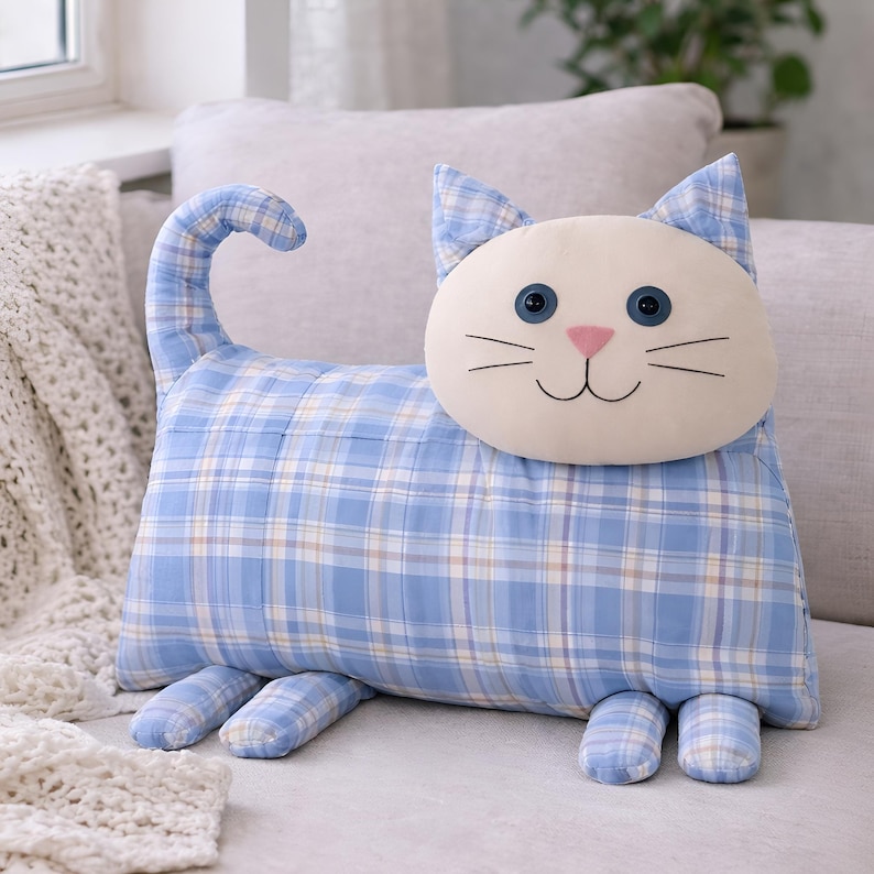 Handmade cat pillow sewing pattern made from blue plaid men’s shirt fabric on a light armchair near window with knitted blanket, beginner DIY plush pillow PDF pattern, upcycled shirt home decor gift idea.