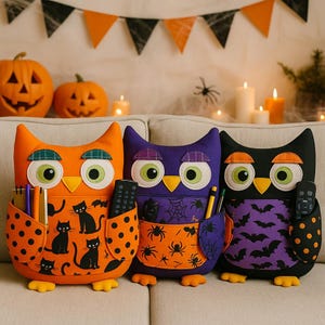 Peut inclure: Trois organisateurs de rangement en forme de hibou sur le thème d'Halloween, orange, violet et noir. Chaque hibou a de grands yeux, une poche et un design unique avec des chats, des araignées et des chauves-souris. Les hiboux sont sur un canapé beige avec des citrouilles et des bougies en arrière-plan.
