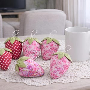 Pink and red fabric strawberry ornaments styled on lace tablecloth with cup in cozy living room