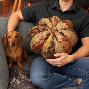 Könnte beinhalten: Ein dekoratives, mehrfarbiges Blumenkissen mit einem braunen, roten und goldenen Paisley-Muster wird von einer Person gehalten. Ein brauner Dackel sitzt neben dem Kissen auf dem Sofa. Das Kissen hat eine runde Form.