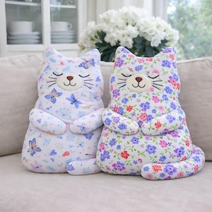 May include: Two plush cat-shaped pillows sitting side by side. The left pillow features a white fabric with a blue and purple butterfly pattern. The right pillow features a white fabric with a dense purple, pink, and red floral pattern.
