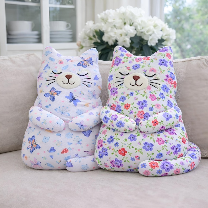 May include: Two plush cat-shaped pillows sitting side by side. The left pillow features a white fabric with a blue and purple butterfly pattern. The right pillow features a white fabric with a dense purple, pink, and red floral pattern.