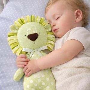Puede incluir: Un peluche de le&oacute;n verde claro con lunares blancos y un lazo es sostenido por un beb&eacute; dormido. El le&oacute;n tiene una nariz marr&oacute;n y una melena verde y amarilla. El beb&eacute; lleva una camiseta blanca y est&aacute; acostado sobre una almohada azul con lunares.