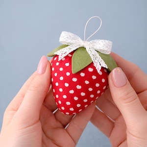 Może przedstawiać: Czerwona ozdoba w kształcie truskawki z białymi kropkami, zielonymi liśćmi z filcu i białą koronkową kokardą. Posiada biały sznurek do zawieszenia. Truskawka trzymana jest w dwóch dłoniach na jasnoniebieskim tle.