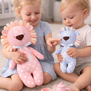 Puede incluir: Dos niños sostienen muñecos de león de peluche. Uno es rosa con lunares blancos, el otro azul con lunares blancos. Un juego de té rosa con tetera, tazas y platillos está en el suelo.