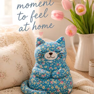 May include: A plush cat toy featuring a bright blue fabric with a colorful floral pattern of small pink, white, and yellow flowers. The cat has embroidered closed eyes, a brown nose, and whiskers. The text "Take a moment to feel at home" is visible.