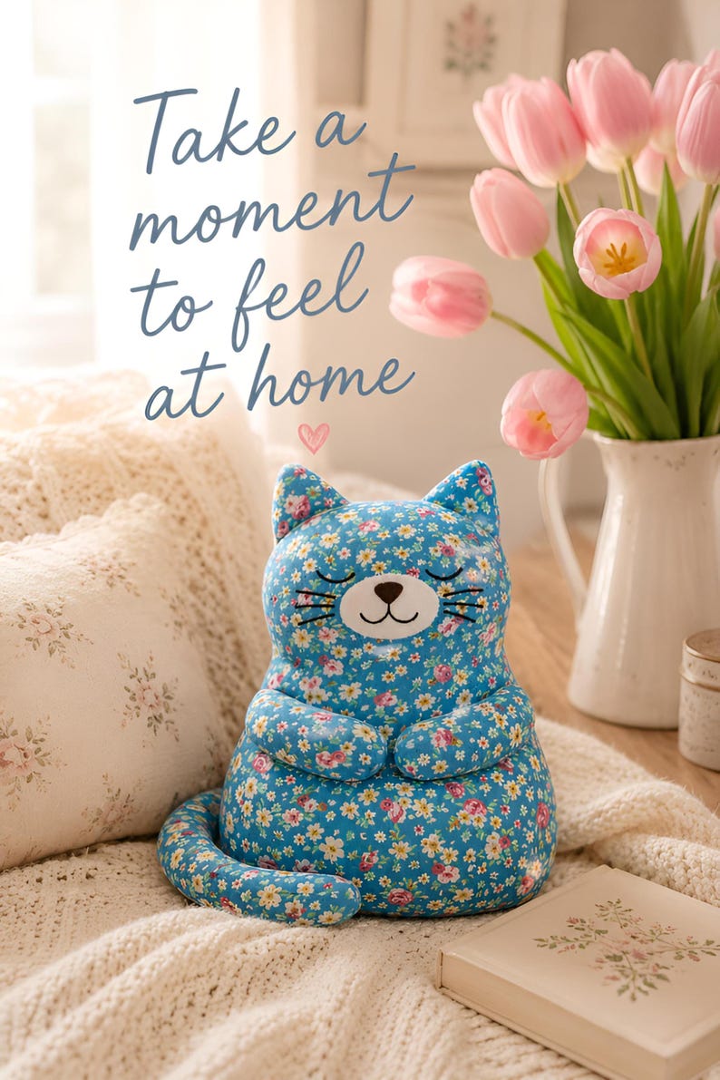 May include: A plush cat toy featuring a bright blue fabric with a colorful floral pattern of small pink, white, and yellow flowers. The cat has embroidered closed eyes, a brown nose, and whiskers. The text "Take a moment to feel at home" is visible.