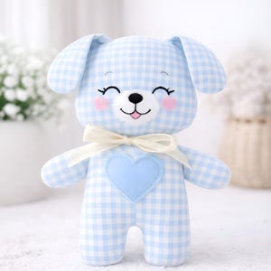 Pode incluir: Um brinquedo de pelúcia em forma de cachorro com um padrão xadrez vichy azul claro e branco. O cachorro tem orelhas caídas, um coração azul claro no peito e um laço creme. O rosto do cachorro tem detalhes bordados, incluindo uma expressão sorridente e bochechas rosadas.