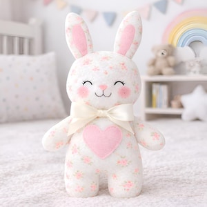 Puede incluir: Un peluche de conejito con un estampado floral blanco y un corazón rosa en el pecho. El conejito tiene orejas internas rosas, mejillas sonrosadas y un lazo color crema. Está sobre una superficie blanca.