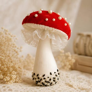 Peut inclure: Un ornement champignon décoratif avec un chapeau en velours rouge vif orné de perles blanches. Le pied est en tissu blanc avec une base décorée de perles noires et blanches. Un volant en dentelle blanche se trouve sous le chapeau.