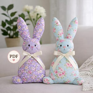 Peut inclure: Deux lapins décoratifs en tissu. L'un est lavande avec un motif floral, l'autre est bleu clair avec un motif floral. Les deux ont des nœuds crème et des visages brodés. Les lapins sont assis droits.