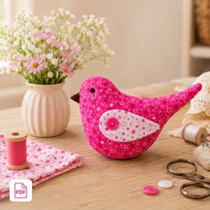Puede incluir: Un pájaro de tela rosa hecho a mano con un ala blanca estampada con estrellas y un pico marrón. El pájaro está decorado con un botón rosa y se encuentra sobre una superficie de madera. También hay un jarrón con flores, carretes de hilo y utensilios de costura.