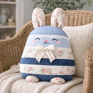 Könnte beinhalten: Ein plüschiges, hasenförmiges Kissen mit einem blauen Jeans-Körper und floralen Akzenten. Der Hase hat lange Ohren mit einem floralen Muster, ein lächelndes Gesicht und eine weiße Spitzenschleife. Das Kissen liegt auf einer weißen Decke.