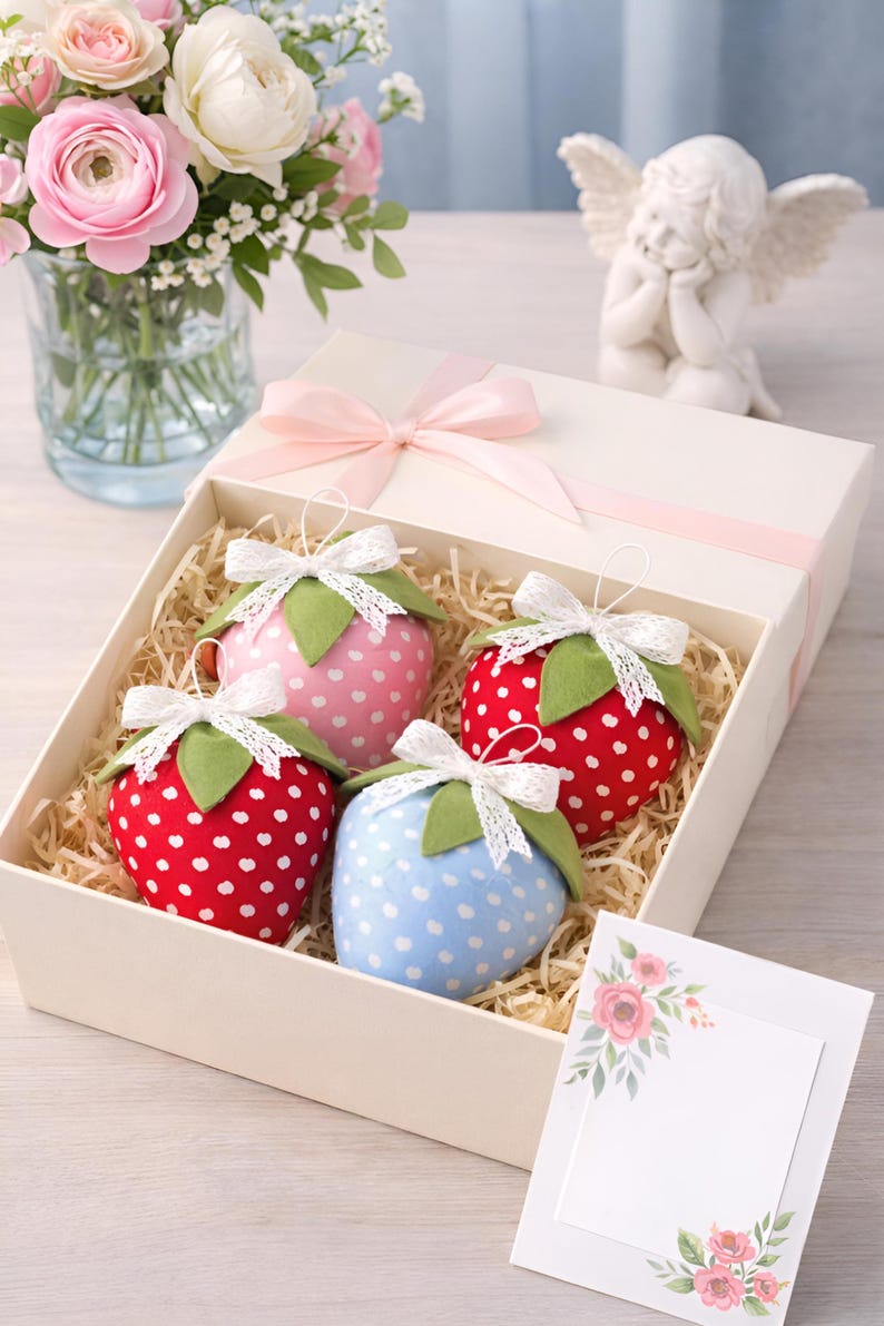 Fabric strawberry sewing pattern gift box with handmade plush berries in different colors, lace bows and greeting card on table with flowers