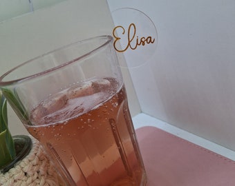 May include: A clear acrylic drink marker with the name "Elisa" written in gold lettering. The marker is placed on the rim of a glass filled with a pink beverage.