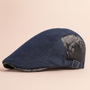 Puede incluir: Gorra de estilo newsboy azul marino con tejido texturizado. La gorra presenta un acento oscuro y estampado en el lateral y un pequeño detalle de hebilla. El borde es de un tono azul más oscuro.