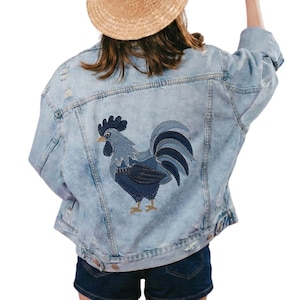 Puede incluir: Una chaqueta de mezclilla azul claro con efecto lavado al ácido que presenta un gran gallo bordado en la espalda. El gallo está representado en tonos azul oscuro y azul claro con pico y patas amarillas. La chaqueta tiene mangas largas.