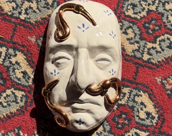 May include: A ceramic wall mask featuring a cream-colored face with small blue floral accents. The mask is adorned with three metallic gold-toned, curved, worm-like sculptures that wrap around the forehead, left cheek, and nose area.