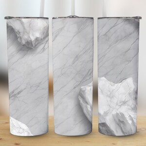 Op de afbeelding: Drie cilindrische bekers met een wit en grijs marmerpatroon, compleet met deksels en rietjes. Het ontwerp bevat witte rotsformaties. De bekers staan op een houten oppervlak.