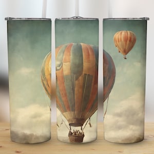 May include: Three stainless steel tumblers with a vintage-inspired design featuring hot air balloons against a cloudy sky. The tumblers have a white background with a faded blue sky and clouds. The hot air balloons are in shades of blue, red, and brown.