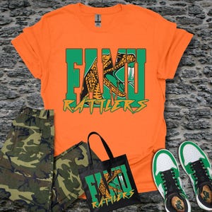 May include: An orange short-sleeved t-shirt featuring a green and orange graphic of a snake and the text FAMU RATTLERS. Also included is a black tote bag with the same snake graphic and text, camouflage pants, and white and green sneakers.