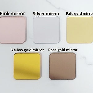 Puede incluir: Cinco espejos cuadrados de diferentes colores: rosa, plata, oro p&aacute;lido, oro amarillo y oro rosa. Los espejos est&aacute;n dispuestos en dos filas sobre una superficie blanca.
