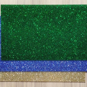 May include: Three rectangular sheets of glitter paper in green, blue, and gold. The glitter is evenly distributed across the surfaces, creating a sparkling effect. The sheets are layered, with the gold sheet at the bottom, followed by blue, and green on top.