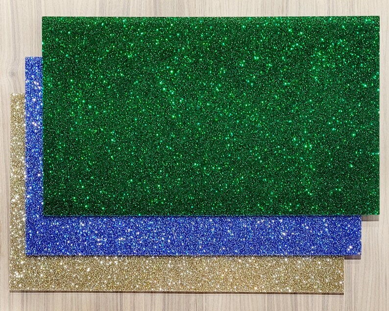 May include: Three rectangular sheets of glitter paper in green, blue, and gold. The glitter is evenly distributed across the surfaces, creating a sparkling effect. The sheets are layered, with the gold sheet at the bottom, followed by blue, and green on top.