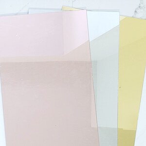 Puede incluir: Cuatro hojas de papel decorativo en diferentes colores. Las hojas est&aacute;n apiladas una encima de la otra. La hoja superior es rosa, la segunda hoja es blanca, la tercera hoja es gris claro y la hoja inferior es dorada.