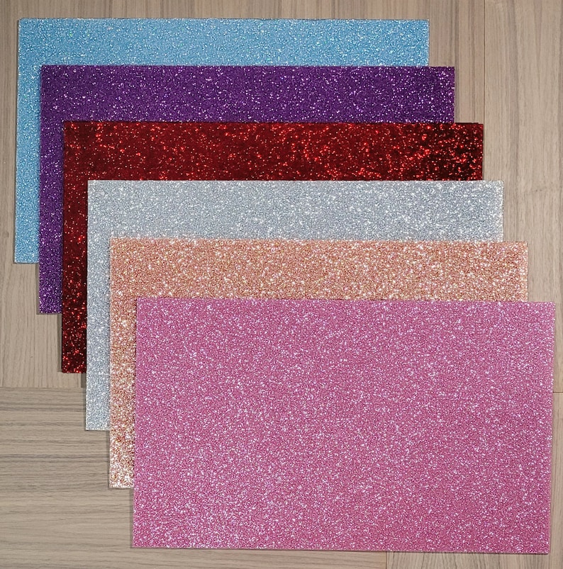 May include: A collection of rectangular glitter sheets in various colors, including light blue, purple, red, silver, rose gold, and pink. The sheets are arranged in a layered fan pattern, showcasing the sparkling texture. The background is a light wood grain.
