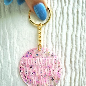 May include: Pink glitter keychain with a gold chain and ring. The keychain has the text "To live for the hope of it all" in white lettering.