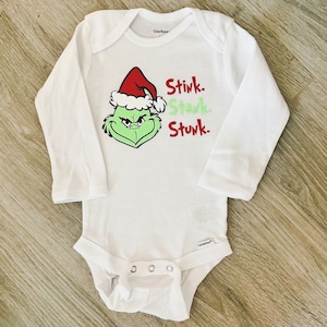 May include: White long-sleeved baby onesie with a green Grinch face wearing a red Santa hat. The text "Stink. Stank. Stunk." is printed in red on the onesie.