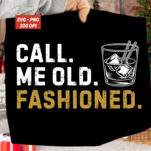 May include: Black t-shirt with the text "CALL. ME OLD. FASHIONED." in white and gold lettering. A graphic of a cocktail glass with ice cubes and straws is on the right side. The shirt is held up by a person.
