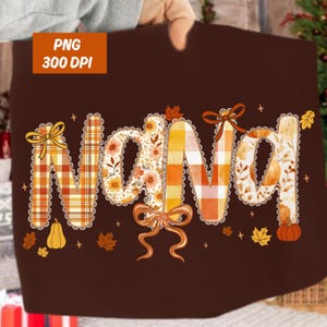 May include: A brown blanket with the word "Nana" in a decorative font. The letters are filled with autumn patterns, including plaid, floral, and leaf designs. The blanket is decorated with pumpkins, leaves, and bows.