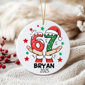 Könnte beinhalten: Weißer Keramik-Weihnachtsschmuck mit goldenem Band. Der Schmuck zeigt die Zahlen "67" in Rot und Grün, jeweils mit einem Weihnachtsmütze. Der Name "BRYAN" und das Jahr "2025" sind darunter mit roten und grünen Sternen aufgedruckt.