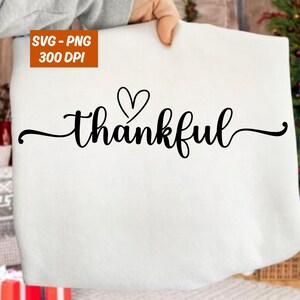May include: A white surface with the word "thankful" in a black, cursive font, with a heart above the "a". The image also includes the text "SVG - PNG 300 DPI" in an orange box.