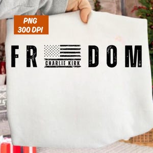 Pode incluir: Camiseta branca com a palavra "FREEDOM" em fonte grande, preta e desgastada. O design incorpora a bandeira americana, com o texto "CHARLIE KIRK" abaixo. A camiseta é segurada por uma pessoa.