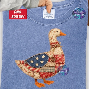 May include: A light blue t-shirt with a patchwork goose design in red, white, and blue. The goose features stars and stripes. The shirt has a crew neck and a tag with the text "CL COLOURS".