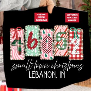Puede incluir: Camiseta negra con un diseño navideño. Muestra los números 46052 en un estilo festivo, incorporando patrones como cuadros escoceses y bastones de caramelo. El texto "small town christmas LEBANON, IN" está escrito debajo.