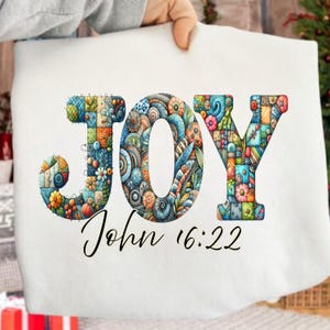 Può includere: Tessuto bianco con la parola "JOY" in lettere colorate e decorate. Le lettere sono riempite con disegni intricati e motivi floreali. Sotto "JOY" c'è il testo "John 16:22" in corsivo.