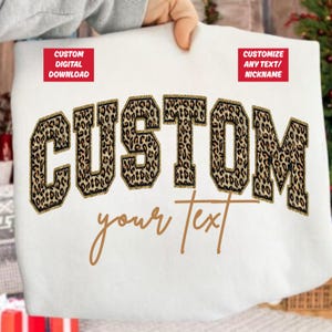 May include: White sweatshirt featuring the word "CUSTOM" in large leopard print letters with gold glitter edging. The words "your text" are written in a cursive font below. Two red rectangles with white text are in the top corners.