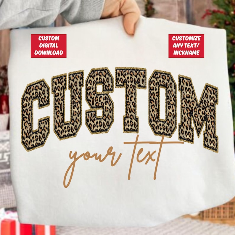 May include: White sweatshirt featuring the word "CUSTOM" in large leopard print letters with gold glitter edging. The words "your text" are written in a cursive font below. Two red rectangles with white text are in the top corners.