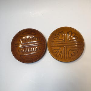 May include: Two round, brown ceramic dishes with raised designs. One features a series of curved lines, while the other has a cross-like pattern with leaf-like shapes. The dishes have a glossy finish.