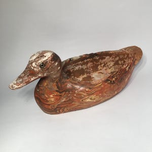 May include: A wooden duck decoy with a weathered, marbled appearance. The duck features a white and brown head, a long beak, and a body with shades of brown, orange, and cream. The decoy is designed to float on water and attract real ducks.