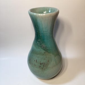 May include: A ceramic vase with a flared rim and a rounded base, featuring a glossy glaze that transitions from a pale cream at the top to a deep teal and seafoam green, with subtle brown speckling and mottled textures throughout the surface.