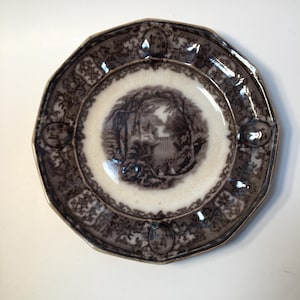May include: An antique octagonal plate with a sepia-toned landscape scene in the center. The plate features a decorative border with floral and geometric patterns. The rim has a scalloped edge, adding to its vintage charm.