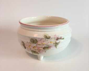 May include: A white ceramic planter with a pink rim and floral design. The planter has three legs and is perfect for displaying small plants.