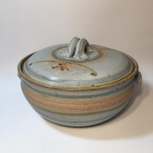 May include: A round ceramic casserole dish with a matching lid featuring a light blue and tan glaze with subtle brown speckles. The lid has a central handle, and the base includes two side handles, all finished in a smooth, mottled stoneware texture.
